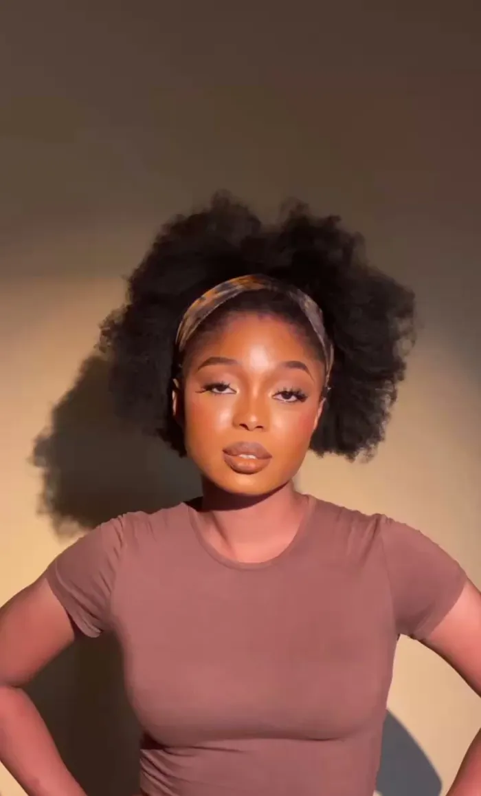 Angela with a natural afro and headband in a brown fitted tee, hands on hips