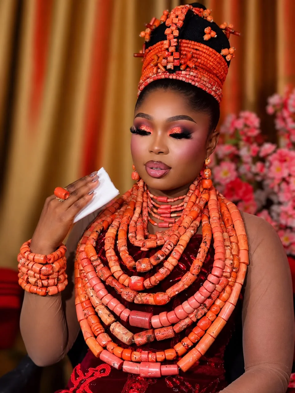 Angela in a full Edo coral bead regalia: a tiered coral crown, layered coral necklaces and stacked coral bracelets over deep red velvet, a tissue in her raised hand, against a gold curtain and pink blossom