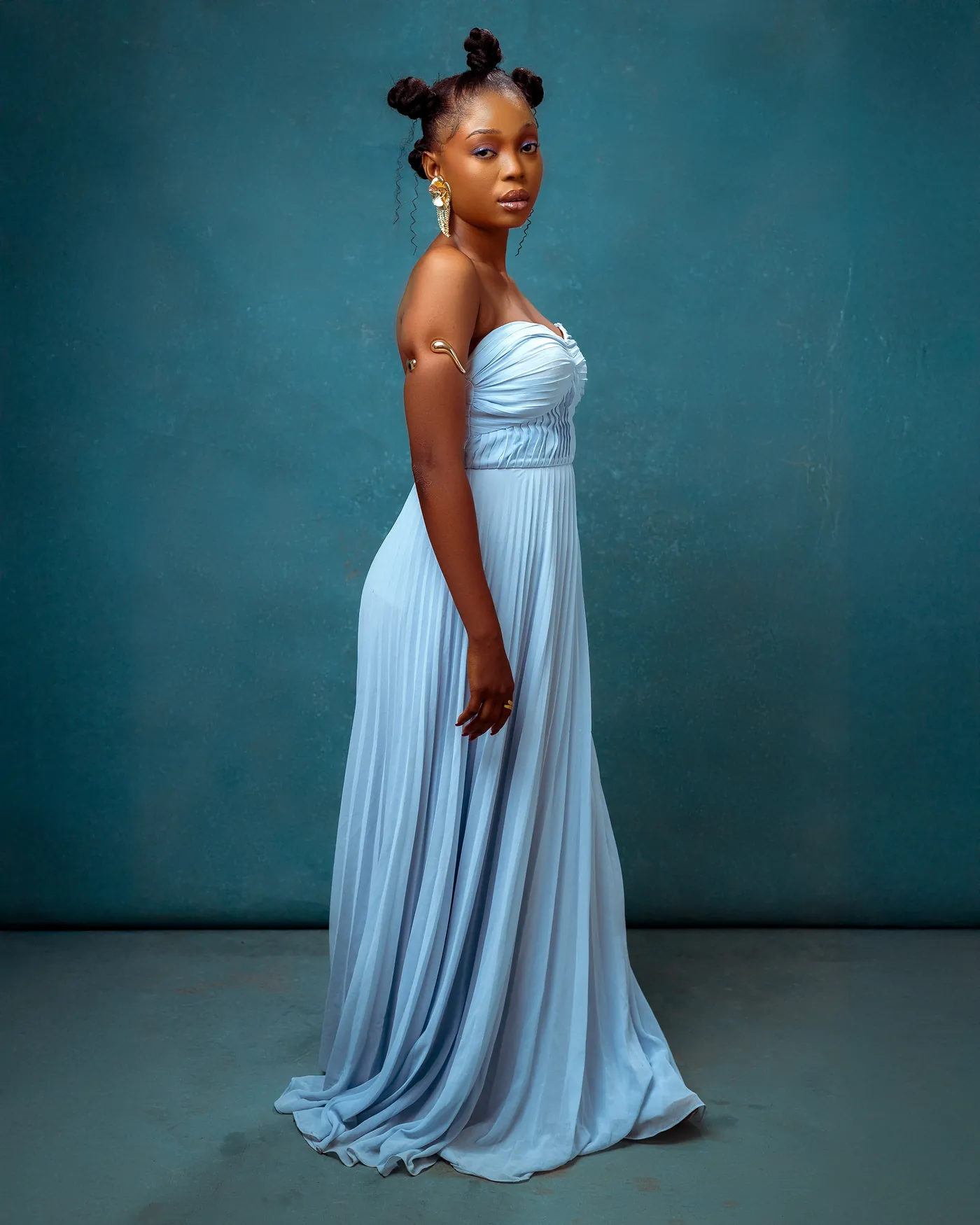 Angela standing full length in a long pale blue pleated gown against a teal studio wall