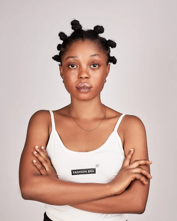 Digital: Angela standing against a plain grey wall in a white vest, arms crossed, hair in bantu knots