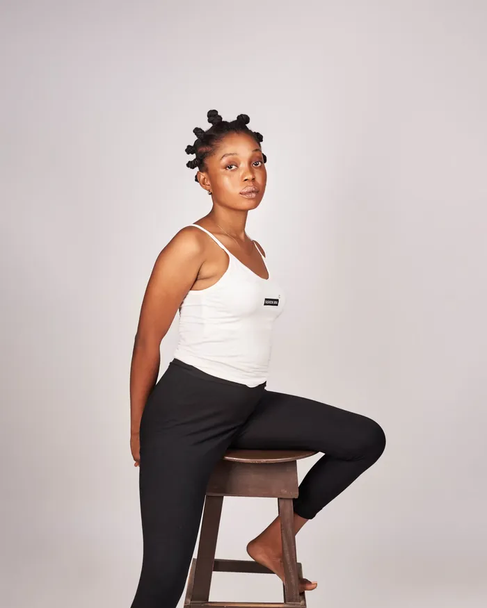 Digital: Angela seated on a wooden stool in a white vest and black trousers against a plain grey wall