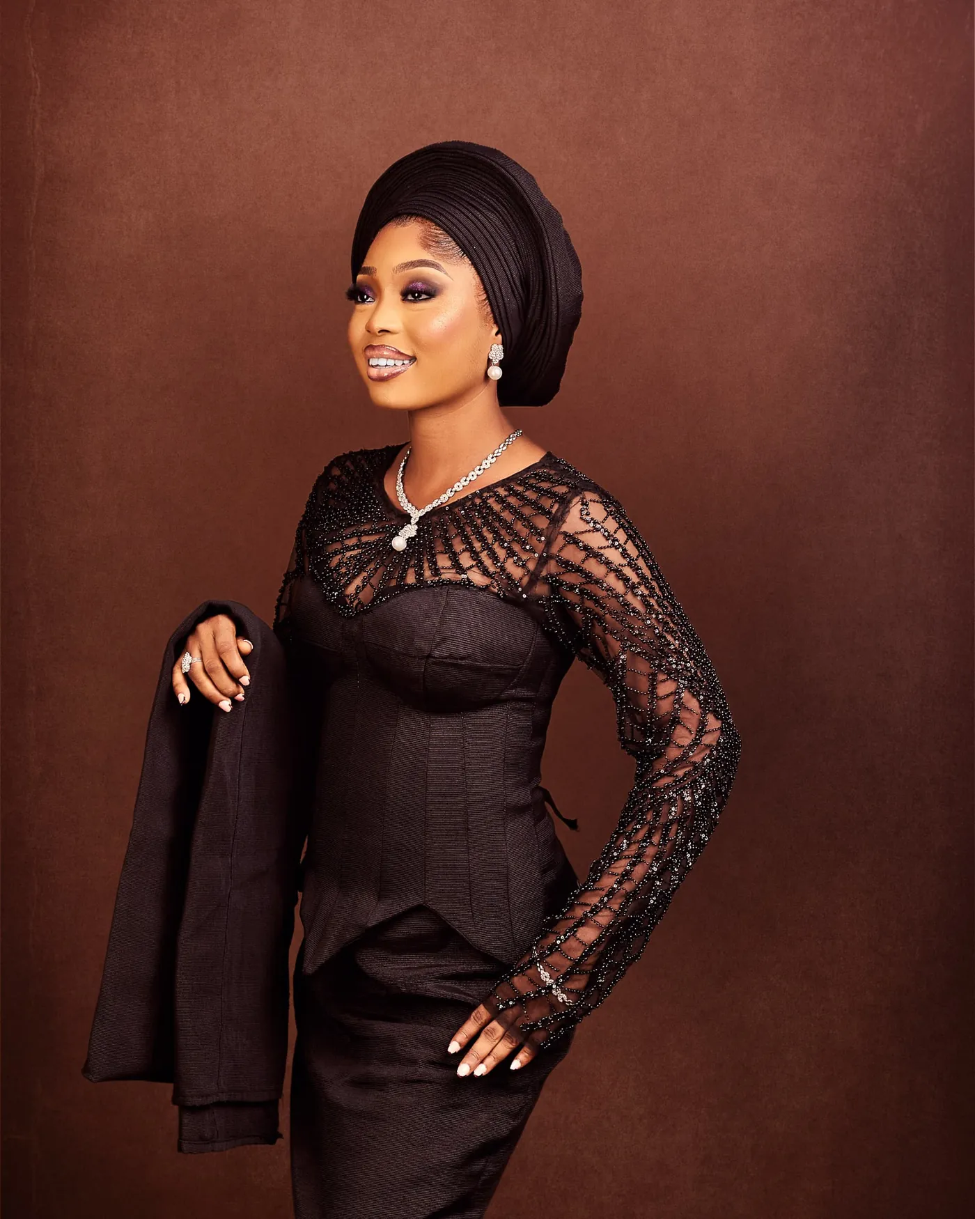 Angela in a black beaded lace top and matching gele, hand on hip, against a brown backdrop