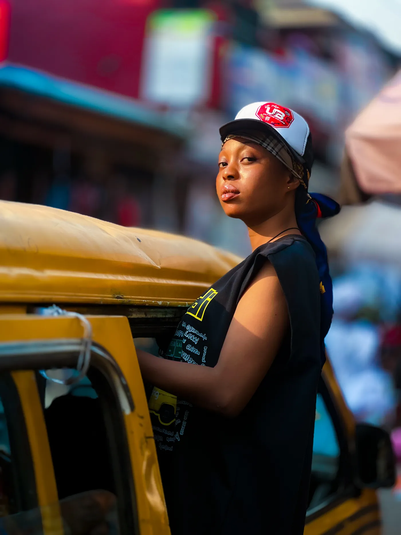 Angela in a trucker cap and sleeveless black tee beside a yellow Lagos danfo bus on a busy street