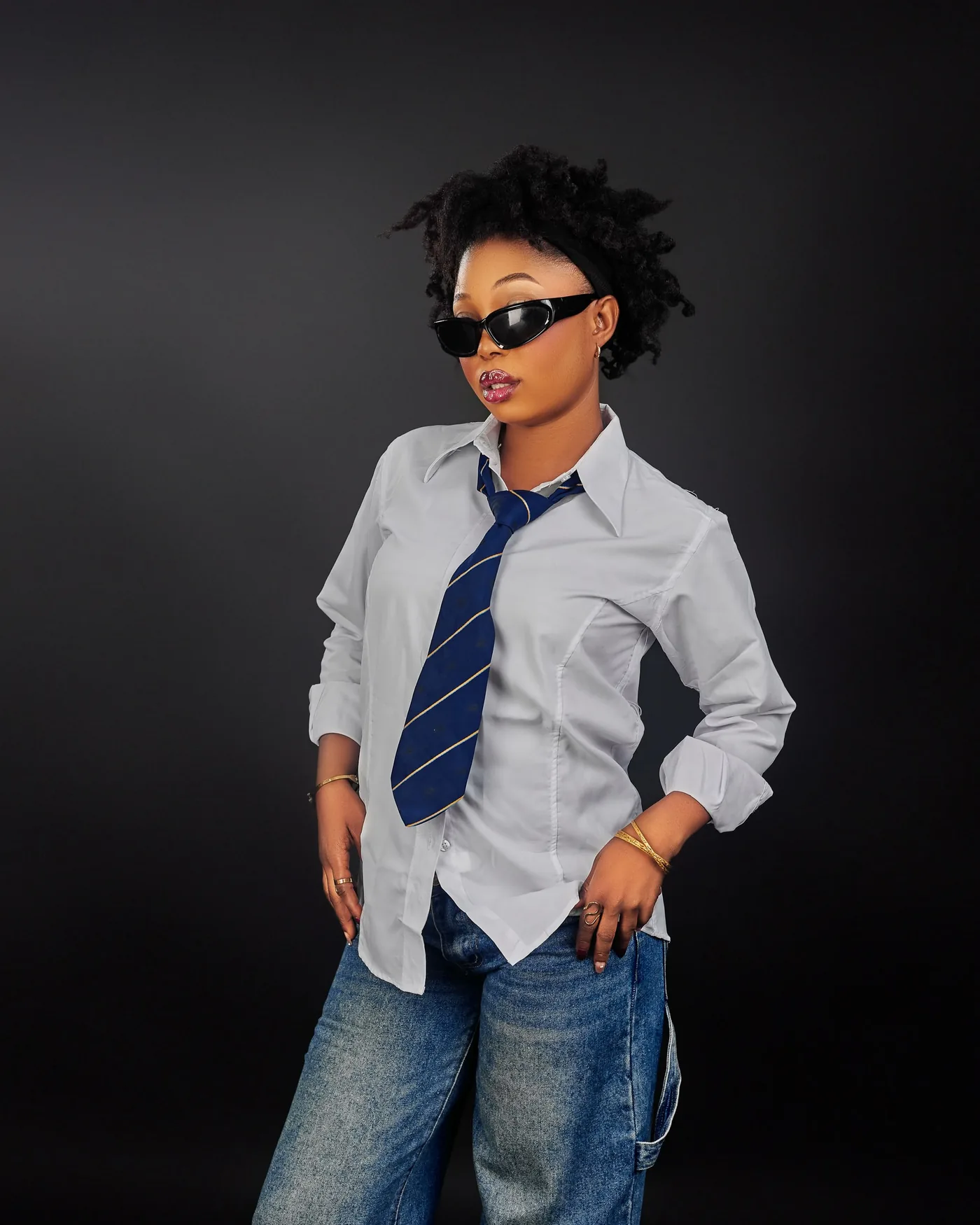 Angela in wide black sunglasses, a white shirt with a loose navy and gold striped tie and wide jeans, a hand on each hip, against a dark studio backdrop
