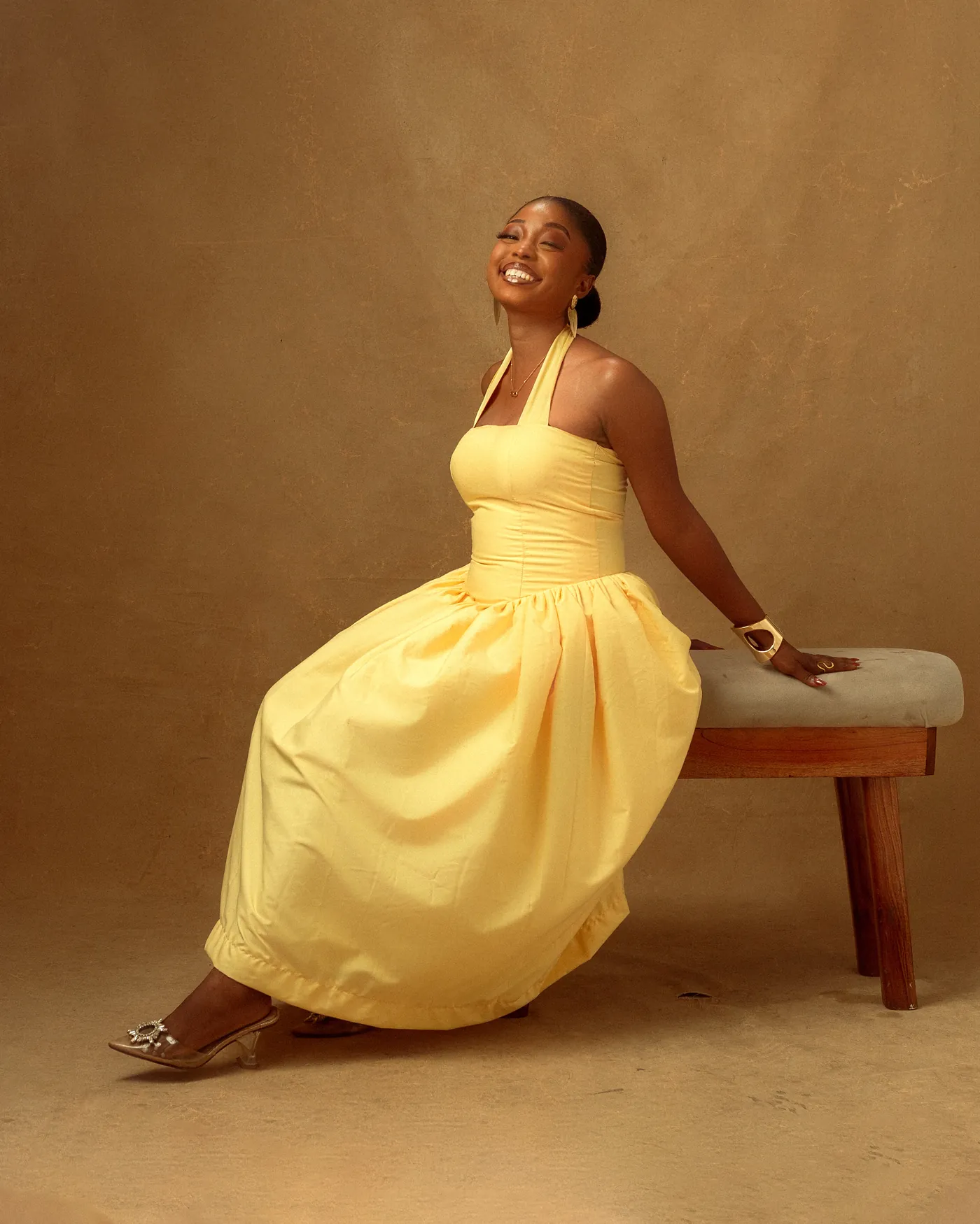 Angela seated on a bench in a pale yellow halter dress, looking up, against a warm sand backdrop